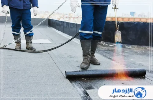 عاملان يثبتان العزل المائي بالسقف باستخدام موقد لهب لتسخين المادة اللاصقة.